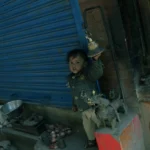 A young child standing beside a blue shuttered shop, reaching for a hanging bell, symbolizing the importance of tax and rights in addressing poverty.
