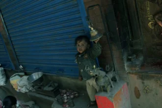 A young child standing beside a blue shuttered shop, reaching for a hanging bell, symbolizing the importance of tax and rights in addressing poverty.