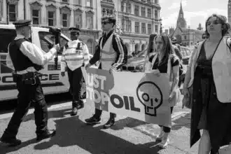 El escenario involucra a un oficial de policía filmando a un activista de Just Stop Oil en Whitehall, cerca de Parliament Square, el 20 de mayo de 2023. Esta situación ejemplifica la compleja dinámica entre violencia y no violencia, a menudo denominada "Binario Violencia-No Violencia", dentro del contexto del activismo social y la aplicación de la ley.