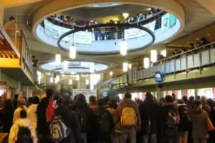 Photo of a bustling anti-racism protest inside the York University student center. Students from diverse backgrounds gather en masse, filling the multi-level atrium. They stand attentively, some with backpacks on, facing a central point of interest, possibly a speaker or focal event. Banners drape over the railings of the upper floors, with messages promoting equality and denouncing racism. The image captures a moment of peaceful yet powerful advocacy, symbolizing a collective quest for expression and equity in an academic setting.