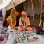 A Naga Sadhu and other Sadhus in traditional saffron attire embody a secular balance, meditating and discussing spirituality amidst simple earthly possessions at a religious camp in India.