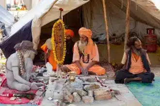 A Naga Sadhu and other Sadhus in traditional saffron attire embody a secular balance, meditating and discussing spirituality amidst simple earthly possessions at a religious camp in India.