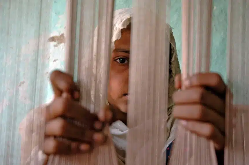 A young girl is peering through white threads, presumably of a loom, in a room with a weathered teal wall. Her expression is solemn, and she holds the threads with her hands, indicating her involvement in labor as a child weaver. This image encapsulates the harrowing realities of modern slavery within the carpet weaving industry in Lahore's district. Her youthful appearance and the setting suggest her role is not by choice but due to the coercive conditions of child labor, a form of modern slavery.
