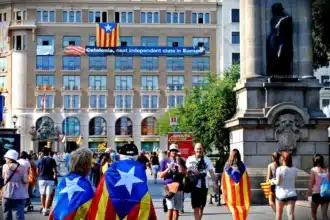 Une foule diversifiée manifestant en faveur de l'indépendance catalane, avec des drapeaux colorés et une banderole promouvant la Catalogne comme un futur État indépendant, mettant en avant la diversité nationale dans un contexte urbain européen.