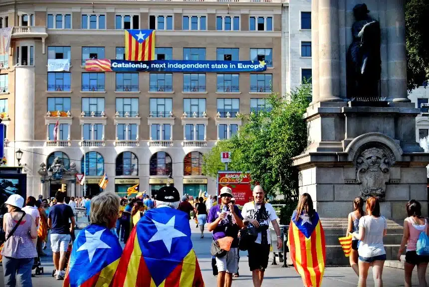 Une foule diversifiée manifestant en faveur de l'indépendance catalane, avec des drapeaux colorés et une banderole promouvant la Catalogne comme un futur État indépendant, mettant en avant la diversité nationale dans un contexte urbain européen.