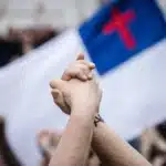 L'image montre un gros plan de deux mains jointes en signe de solidarité ou d'unité. En arrière-plan, il y a un drapeau flou comportant une croix, qui peut symboliser le nationalisme chrétien, une idéologie politique qui cherche à fusionner les identités chrétienne et nationale. Le drapeau semble prendre une place centrale dans un rassemblement, suggérant une manifestation publique ou un événement politique où les symboles chrétiens sont utilisés dans un contexte nationaliste.
