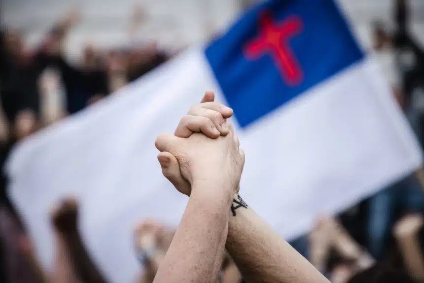 The image displays a close-up of two hands clasped together in solidarity or unity. In the background, there is a blurred flag featuring a cross, which may symbolize Christian nationalism, a political ideology that seeks to merge Christian and national identities. The flag seems to take center stage in a gathering, suggesting a public demonstration or political event where Christian symbols are being used in a nationalistic context.