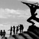 The New Man statue in Durres, Albania, stands as a silhouette against the sky, symbolizing strength and forward-looking determination, while a group of lively people mirror its stance, celebrating the spirit of unity and progress.