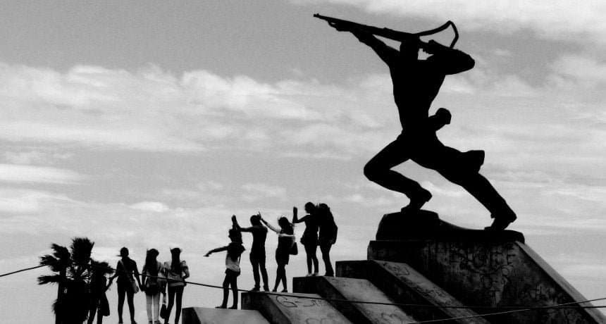 The New Man statue in Durres, Albania, stands as a silhouette against the sky, symbolizing strength and forward-looking determination, while a group of lively people mirror its stance, celebrating the spirit of unity and progress.