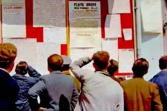 A group of people is gathered around a public bulletin board under the public scrutiny of Albania's communist regime. The board displays documents called "fletërrufe" or "lightning sheets," which contain denunciations or critiques. Individuals are intently reading or discussing the content of these papers.