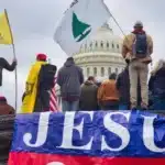 Un grupo de individuos con banderas y pancartas, una de ellas destacando prominentemente el nombre de "JESÚS", parados frente a un edificio significativo que parece ser el Capitolio de EE. UU. Esta escena podría estar asociada con los eventos del 6 de enero de 2021, donde la intersección del activismo político, el fervor religioso y el sentimiento nacionalista estaban visiblemente en exhibición. La presencia de simbolismo religioso, como la pancarta de "JESÚS", sugiere la involucración de ciertos grupos que pueden mezclar sus puntos de vista políticos con sus creencias religiosas, lo que se alinea con ideologías como las propagadas por el movimiento de Identidad Cristiana.