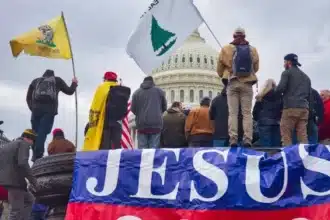 A group of individuals with flags and banners, one prominently featuring the name "JESUS", standing before a significant building that appears to be the U.S. Capitol. This scene could be associated with the events of January 6, 2021, where the intersection of political activism, religious fervor, and nationalist sentiment were visibly on display. The presence of religious symbolism, such as the "JESUS" banner, suggests the involvement of certain groups that may blend their political views with their religious beliefs, which aligns with ideologies like those propagated by the Christian Identity movement.