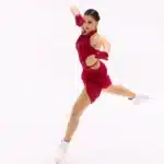 L'image montre Kamila Valieva habillée d'un costume rouge, se produisant sur la glace avec une posture posée et gracieuse. Elle porte des gants rouges assortis et des patins blancs. Son expression concentrée et l'athlétisme affiché suggèrent qu'elle est en plein milieu d'une routine compétitive, démontrant l'art et la précision du patinage artistique.