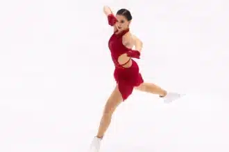 L'image montre Kamila Valieva habillée d'un costume rouge, se produisant sur la glace avec une posture posée et gracieuse. Elle porte des gants rouges assortis et des patins blancs. Son expression concentrée et l'athlétisme affiché suggèrent qu'elle est en plein milieu d'une routine compétitive, démontrant l'art et la précision du patinage artistique.
