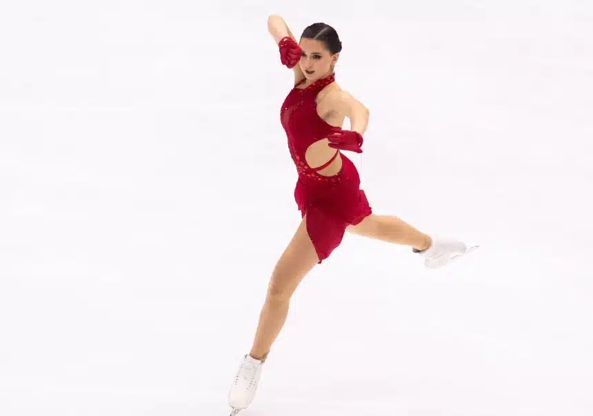 The image shows Kamila Valieva dressed in a red costume, performing on ice with a poised and graceful stance. She wears matching red gloves and white skates. Her focused expression and the athleticism displayed suggest she is in the midst of a competitive routine, demonstrating the artistry and precision of figure skating.