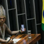 In the Senate Chamber of Brazil, a special session is held to honor indigenous peoples. At the table, Chief Xucuru, Marcos Xukuru records the session using his cellphone.