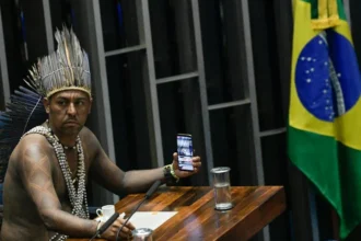 In the Senate Chamber of Brazil, a special session is held to honor indigenous peoples. At the table, Chief Xucuru, Marcos Xukuru records the session using his cellphone.