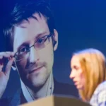 The image shows a woman speaking at a podium during an event, with a large projected image of Edward Snowden in the background. Snowden is adjusting his glasses with one hand and looks directly ahead with a serious expression. The word "traitor" could refer to how some people perceive Snowden's actions of leaking classified NSA information. This act is seen by some as a betrayal of his country, while others view him as a courageous whistleblower who exposed government violations of privacy.