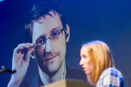 The image shows a woman speaking at a podium during an event, with a large projected image of Edward Snowden in the background. Snowden is adjusting his glasses with one hand and looks directly ahead with a serious expression. The word "traitor" could refer to how some people perceive Snowden's actions of leaking classified NSA information. This act is seen by some as a betrayal of his country, while others view him as a courageous whistleblower who exposed government violations of privacy.
