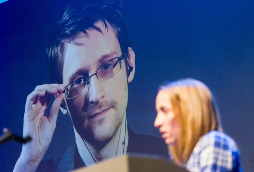 The image shows a woman speaking at a podium during an event, with a large projected image of Edward Snowden in the background. Snowden is adjusting his glasses with one hand and looks directly ahead with a serious expression. The word "traitor" could refer to how some people perceive Snowden's actions of leaking classified NSA information. This act is seen by some as a betrayal of his country, while others view him as a courageous whistleblower who exposed government violations of privacy.