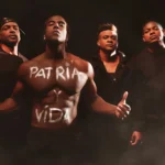 Quatre hommes se tiennent ensemble, dont un au centre affiche les mots "Patria y Vida" écrits sur son torse nu. L'image évoque l'esprit de la chanson "Patria y Vida", qui est devenue un hymne de la résistance contre la dictature cubaine. Leurs expressions sérieuses et leurs vêtements sombres soulignent le message puissant de la chanson et l'unité des Cubains dans leur lutte pour la liberté et la justice.