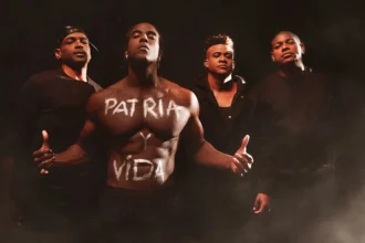 Four men stand together, with one in the center displaying the words "Patria y Vida" written across his bare chest. The image evokes the spirit of the song "Patria y Vida," which has become an anthem of resistance against the Cuban dictatorship. Their serious expressions and dark clothing underline the song's powerful message and the unity of Cubans in their fight for freedom and justice.