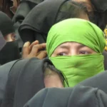A group of Rohingya refugees, with a woman in a green headscarf prominently in the foreground. The image highlights the plight of the Rohingya people, a minority group from Myanmar, many of whom have sought refuge in neighboring countries. They face significant challenges, including limited legal protection and uncertain living conditions.