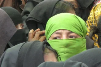A group of Rohingya refugees, with a woman in a green headscarf prominently in the foreground. The image highlights the plight of the Rohingya people, a minority group from Myanmar, many of whom have sought refuge in neighboring countries. They face significant challenges, including limited legal protection and uncertain living conditions.