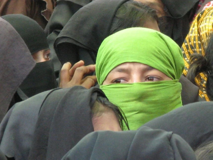 A group of Rohingya refugees, with a woman in a green headscarf prominently in the foreground. The image highlights the plight of the Rohingya people, a minority group from Myanmar, many of whom have sought refuge in neighboring countries. They face significant challenges, including limited legal protection and uncertain living conditions.