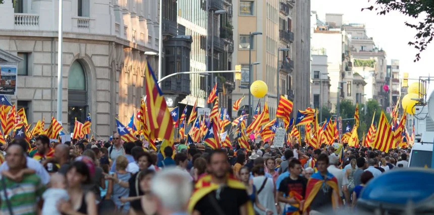 A large crowd of people participating in a Catalonian independence protest, marching through city streets and waving numerous Catalan flags, advocating for political rights and independence from Spain.