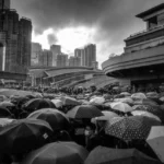 This image captures a significant moment during the 2019 Hong Kong anti-extradition bill protests. It shows a large crowd gathered under a sea of umbrellas, a symbol of resistance, amidst a rainy setting in an urban environment. The towering skyscrapers in the background and curved structures of a nearby transport hub enhance the metropolitan feel of the scene. The photo, taken in black and white, adds a dramatic and somber tone, emphasizing the unity and determination of the protesters. The dense congregation of umbrellas not only protects against the rain but also visually represents the collective spirit of the people standing against the proposed legislation.