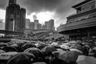 This image captures a significant moment during the 2019 Hong Kong anti-extradition bill protests. It shows a large crowd gathered under a sea of umbrellas, a symbol of resistance, amidst a rainy setting in an urban environment. The towering skyscrapers in the background and curved structures of a nearby transport hub enhance the metropolitan feel of the scene. The photo, taken in black and white, adds a dramatic and somber tone, emphasizing the unity and determination of the protesters. The dense congregation of umbrellas not only protects against the rain but also visually represents the collective spirit of the people standing against the proposed legislation.