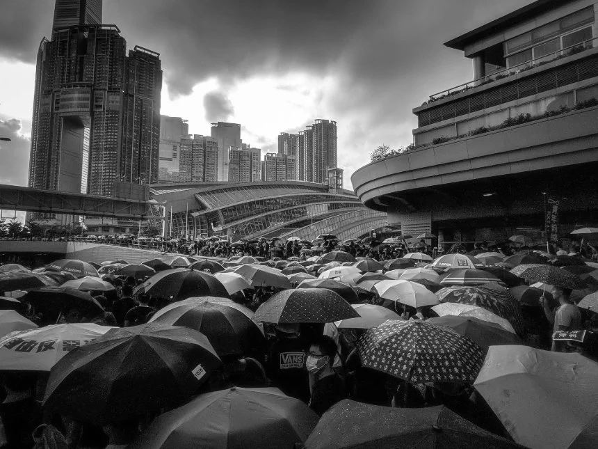 This image captures a significant moment during the 2019 Hong Kong anti-extradition bill protests. It shows a large crowd gathered under a sea of umbrellas, a symbol of resistance, amidst a rainy setting in an urban environment. The towering skyscrapers in the background and curved structures of a nearby transport hub enhance the metropolitan feel of the scene. The photo, taken in black and white, adds a dramatic and somber tone, emphasizing the unity and determination of the protesters. The dense congregation of umbrellas not only protects against the rain but also visually represents the collective spirit of the people standing against the proposed legislation.