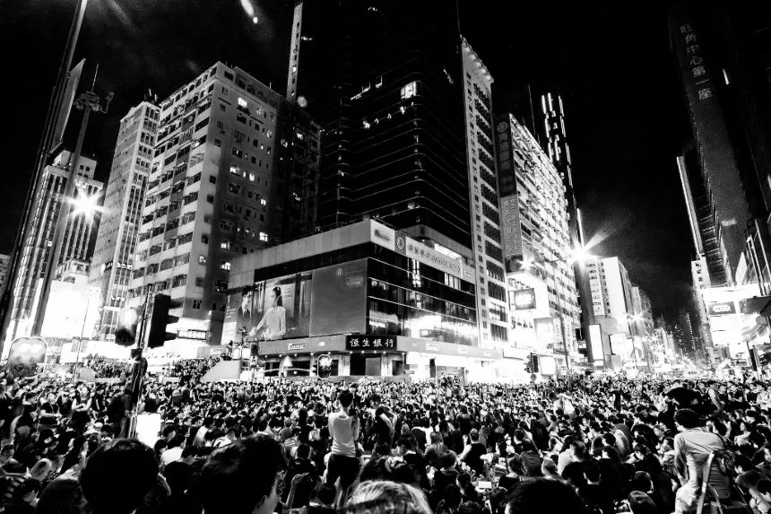 Black and white photo of a massive protest in Hong Kong in 2014, capturing a densely packed crowd of thousands of demonstrators filling a major urban street at night. The protesters are surrounded by towering skyscrapers, lit by bright advertisements and street lights, adding a dramatic effect to the scene. The photo conveys the scale of the protest and the urban environment.