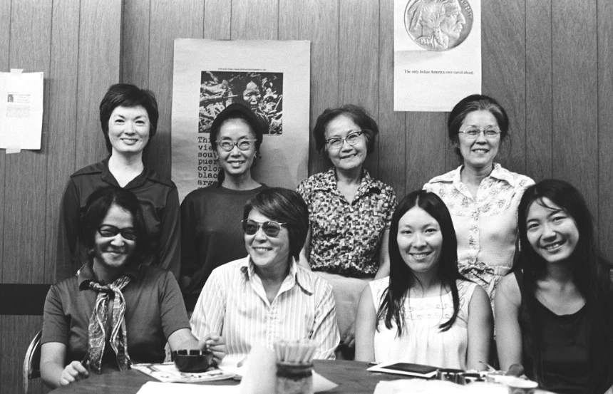 The photo shows several members of Asian Americans for Action along with their guests: Standing from left are guest Elsie Uyematsu [Osajima], Yuri Kochiyama, Minn Matsuda, and Aiko Herzig-Yoshinaga. Seated from left are a feminist from Japan [unnamed], Mitzi Sawada, guest Amy Tachiki [Uyematsu], and Lynn Yamashiro [Taise]. The photo was taken by Mary Uyematsu [Kao] in New York City, 1973, at the United Asians Center, highlighting their activism in race and social justice.