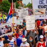 A crowd of Cuban protesters in Canada has gathered to protest against the dictatorship in Cuba. The demonstrators are holding signs expressing their rejection of the communist regime and denouncing the murders and political oppression. Some of the visible signs in the image include messages like "Libertad para los presos políticos" (Freedom for political prisoners), "Patria y Vida" (Homeland and Life), "Viva Cuba libre" (Long live free Cuba), and "We must finish the communism in Cuba." The protest highlights the demand for freedom and human rights for Cubans, as well as the condemnation of the repression and human rights violations by the Cuban government. Additionally, Cuban flags and various images contrasting the situation in Cuba with other realities can be seen.