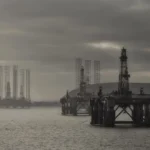 The image displays several oil rigs positioned in a body of water under a cloudy sky that casts a somber, gloomy atmosphere. These massive structures, aligned across the horizon, emphasize the dominant presence of the oil industry amid political instability.