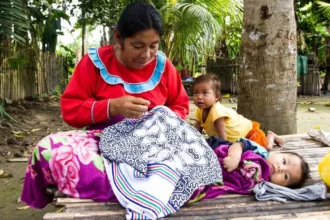 A woman in traditional attire is seated under a tree, meticulously sewing a patterned fabric. Beside her, two young children rest on a wooden platform, surrounded by vibrant blankets and lush greenery, capturing a moment of daily life and vulnerability in a rural setting.