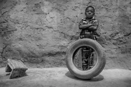 Un niño pequeño está de pie contra una pared texturizada con los brazos cruzados, vistiendo ropa estampada. Frente al niño, hay un neumático grande y en posición vertical. A la izquierda del niño, un pequeño bloque en ángulo descansa en el suelo. La imagen es en blanco y negro, enfatizando el contraste y los detalles de la escena. Esta imagen simboliza la resiliencia y la fuerza de los niños en entornos desafiantes. Sirve como recordatorio de la importancia de abogar y proteger los derechos de los niños, asegurando que tengan acceso a la educación, la seguridad y oportunidades para un futuro mejor.