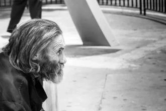 Una foto en blanco y negro que muestra a un anciano sin hogar con una larga barba y ropa desgastada, sentado en una calle de la ciudad. Su expresión es contemplativa mientras observa, representando un recordatorio contundente de la necesidad social de compasión y apoyo para los más vulnerables.