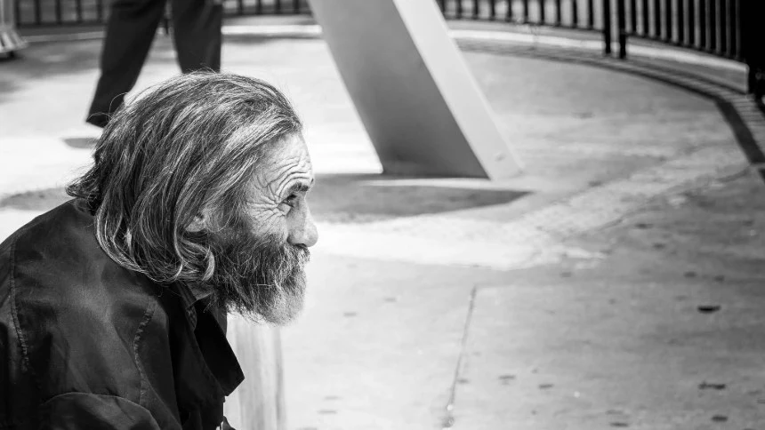 Una foto en blanco y negro que muestra a un anciano sin hogar con una larga barba y ropa desgastada, sentado en una calle de la ciudad. Su expresión es contemplativa mientras observa, representando un recordatorio contundente de la necesidad social de compasión y apoyo para los más vulnerables.