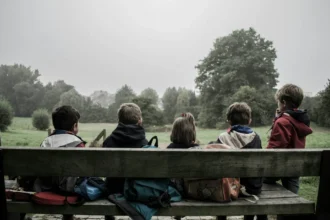 Cinco niños están sentados en un banco de madera al aire libre, mirando hacia un paisaje verde con árboles bajo un cielo nublado. Llevan mochilas y chaquetas, posiblemente durante una actividad o excursión al aire libre. La imagen evoca un momento de paz en la naturaleza, simbolizando la importancia de los derechos de los niños a disfrutar de entornos seguros y saludables, y a tener acceso a oportunidades educativas y recreativas.