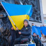 A young boy holds the Ukrainian flag during a demonstration, symbolizing resilience and national pride amidst the ongoing conflict. The crowd in the background showcases strong support for Ukraine.