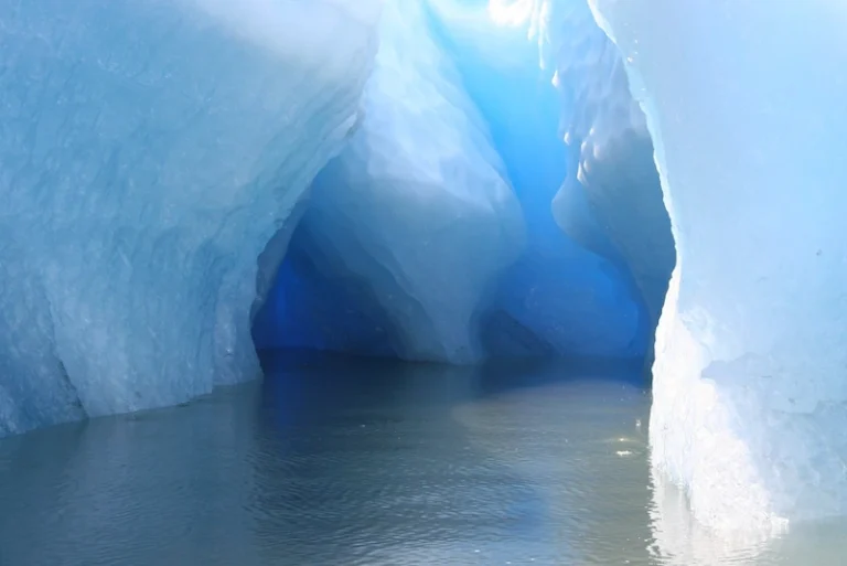 An icy cavern in the Arctic, illuminated by a natural light that reveals a spectrum of blue tones, reflecting off the calm water within.