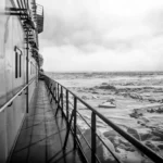 A monochrome view from the deck of an Arctic exploration ship. The photograph captures the ship's side and its extended walkway, leading the eye towards the icy, turbulent Arctic seascape visible in the distance. The image, under a cloudy sky, emphasizes the cold and remote environment of the Arctic region.