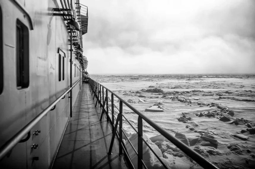 A monochrome view from the deck of an Arctic exploration ship. The photograph captures the ship's side and its extended walkway, leading the eye towards the icy, turbulent Arctic seascape visible in the distance. The image, under a cloudy sky, emphasizes the cold and remote environment of the Arctic region.