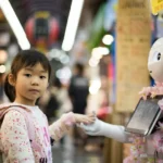 Young child interacting with a humanoid robot adorned with flowers in a busy indoor marketplace, illustrating the integration of artificial intelligence in everyday life.