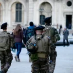 The image shows military personnel patrolling an urban area as part of counterterrorism measures. The soldiers, equipped with tactical gear such as backpacks and communication devices, are walking through a public square. In the background, civilians can be seen near a historic building, illustrating the visible presence of security forces in high-traffic locations to deter potential terrorist threats.