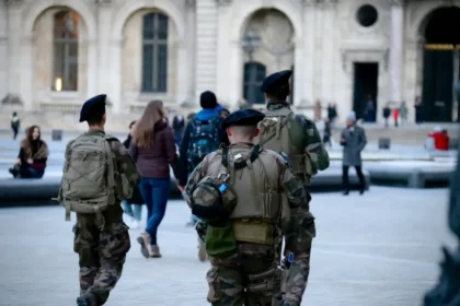 L'image montre du personnel militaire patrouillant dans une zone urbaine dans le cadre de mesures de lutte contre le terrorisme. Les soldats, équipés de matériel tactique tel que des sacs à dos et des dispositifs de communication, marchent sur une place publique. À l'arrière-plan, des civils sont visibles près d'un bâtiment historique, illustrant la présence visible des forces de sécurité dans des lieux très fréquentés pour dissuader les menaces terroristes potentielles.