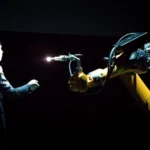 Esta imagen muestra la interacción entre humanos y robots industriales en el lugar de trabajo. Refleja el aumento de la automatización en la producción y el posible desplazamiento laboral que la integración de la inteligencia artificial trae al mercado laboral.